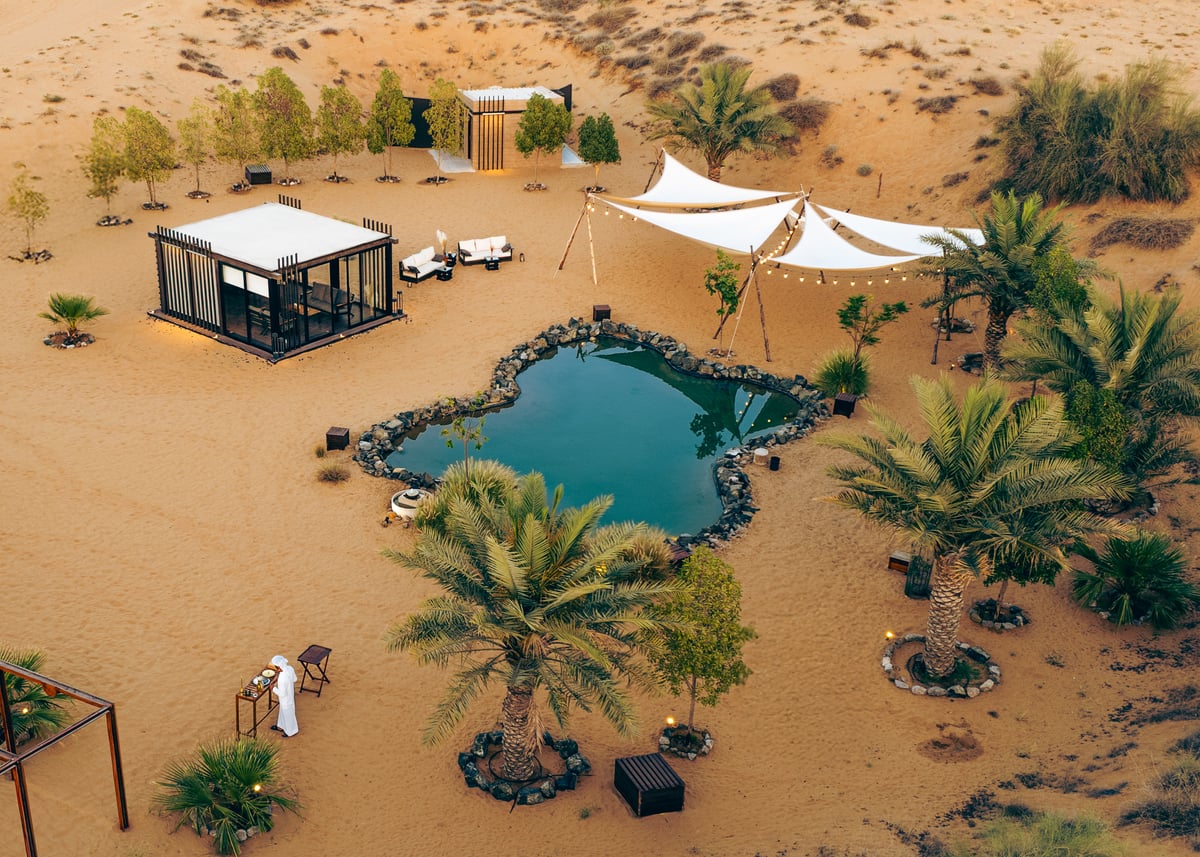 Aerial view of a palm-ringed oasis pool, cabins and shade sails in the dunes