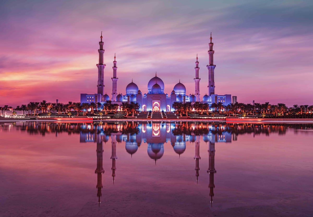 The mosque's minarets and domes glowing violet, mirrored in still water under a pink sky
