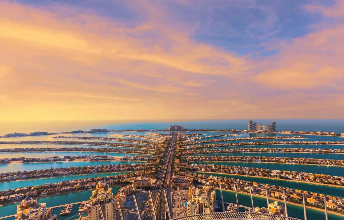 Aerial view of Palm Jumeirah's fronds and trunk at dusk, lit villas under a pink and gold sky
