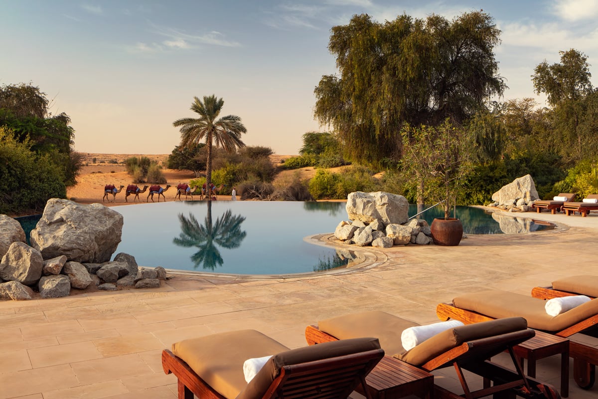 Al Maha main pool edged with rocks and palms, loungers in the foreground and camels walking past on the dunes beyond.