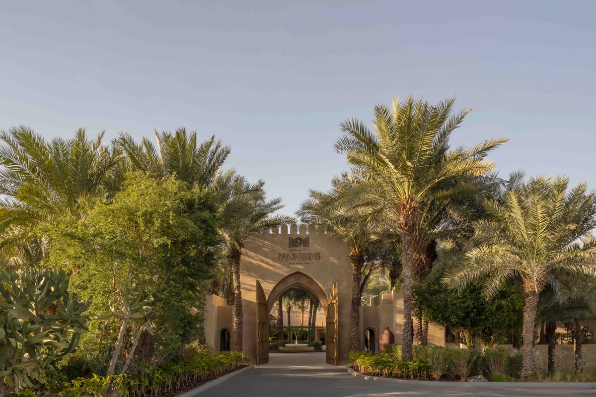 The arched gateway of Bab Al Shams at the end of a paved drive lined with date palms and gardens.