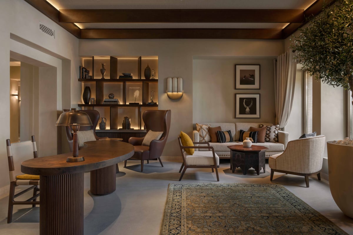 A lounge corner beside reception at Bab Al Shams with armchairs, a round wooden table and lit shelving.