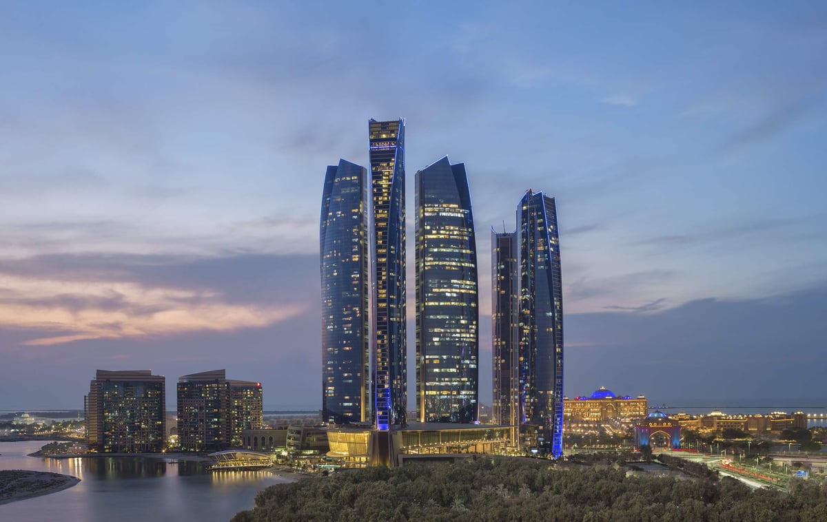 The towers of Conrad Abu Dhabi Etihad Towers lit against a pink and blue dusk sky, with water and low buildings in front.