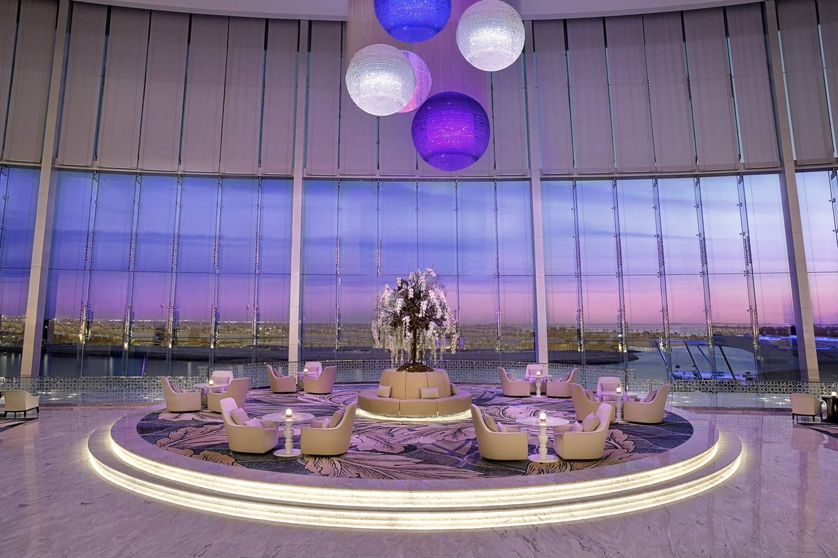 A circular lounge with armchairs around a floral centrepiece under hanging purple glass globes and tall windows at Conrad Abu Dhabi Etihad Towers.