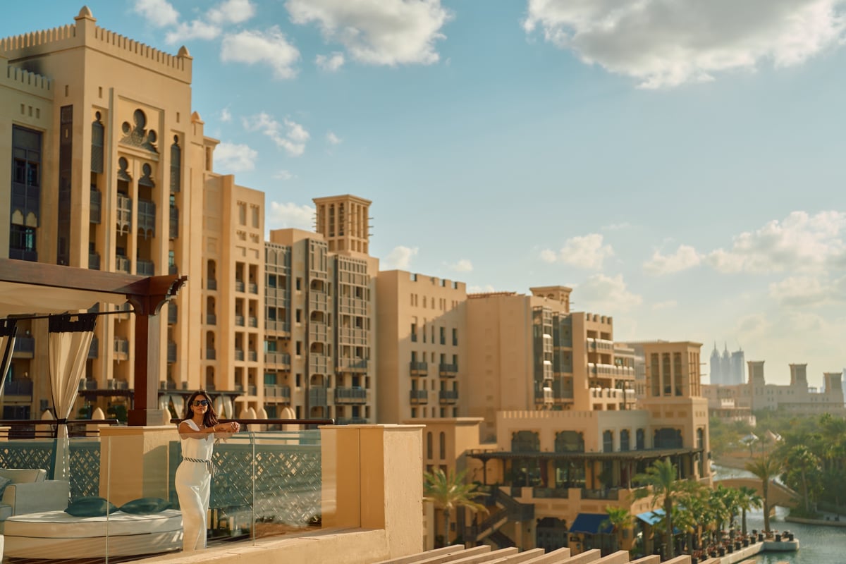 Sandstone balconies and wind towers of Jumeirah Mina A'Salam rise above the waterway, with a woman posing on a terrace.
