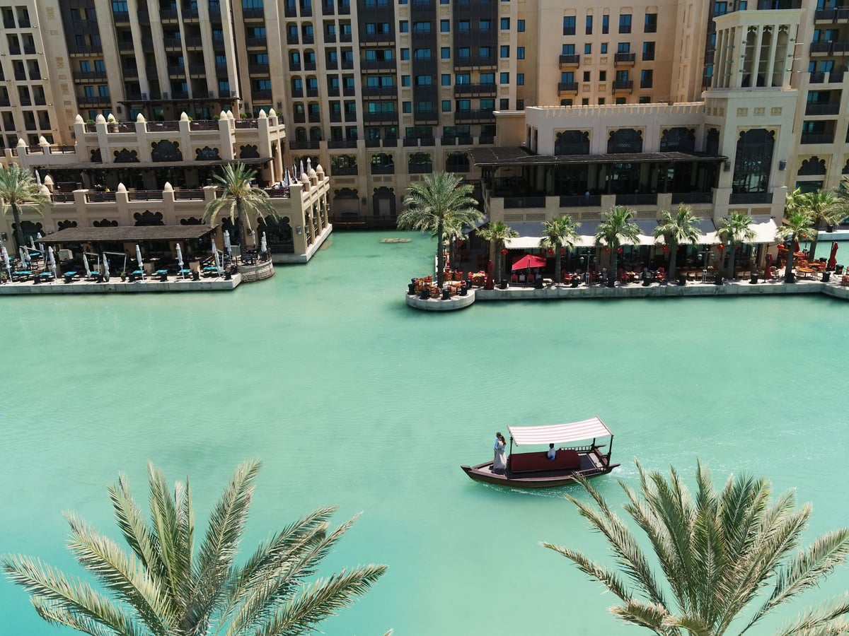 A wooden abra crosses turquoise Madinat Jumeirah waterways below the terraces and towers of Jumeirah Mina A'Salam, framed by palm fronds.