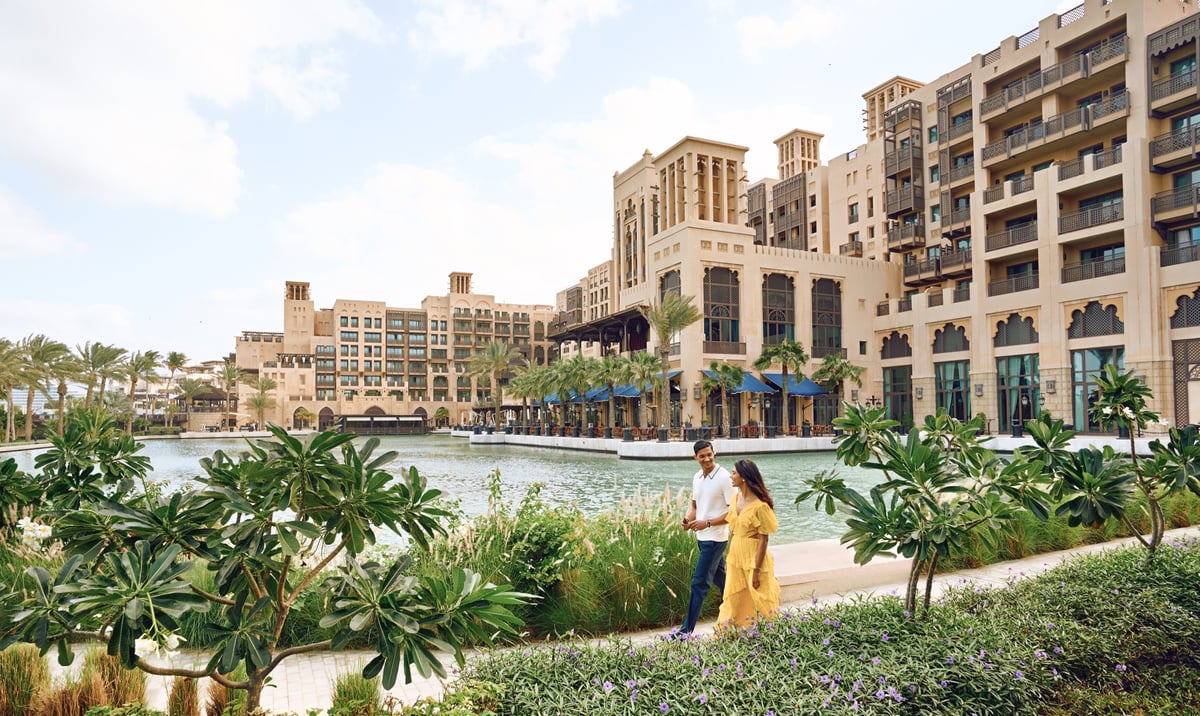 A couple strolls a palm-lined path beside the waterway beneath the tall sandstone wings of Jumeirah Mina A'Salam.