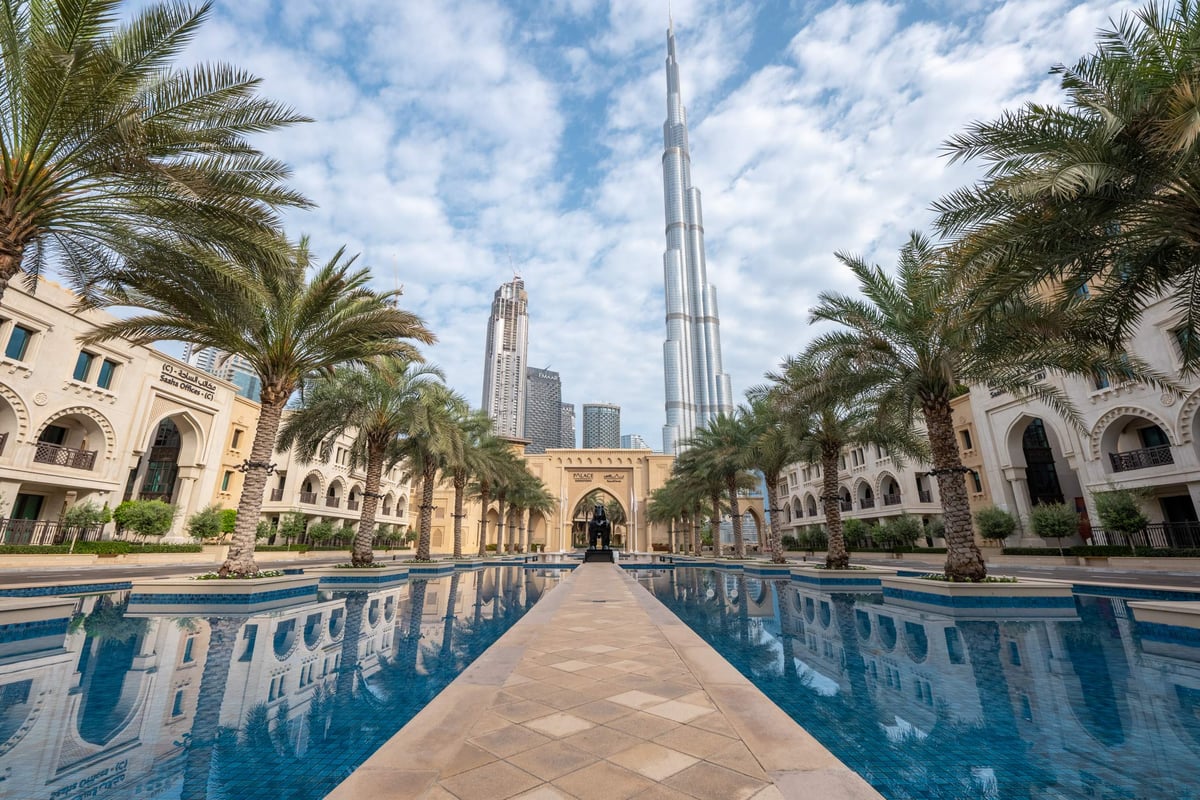 A paved walkway between reflecting pools and palm trees leads to Palace Downtown's arched entrance, with Burj Khalifa rising behind.
