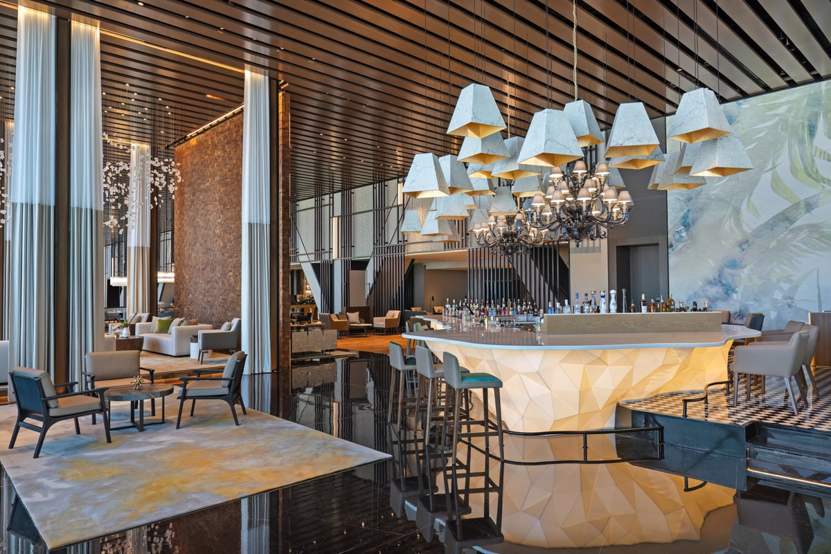 A faceted, glowing bar counter beneath a cluster of white shaded pendants, armchairs set around low tables