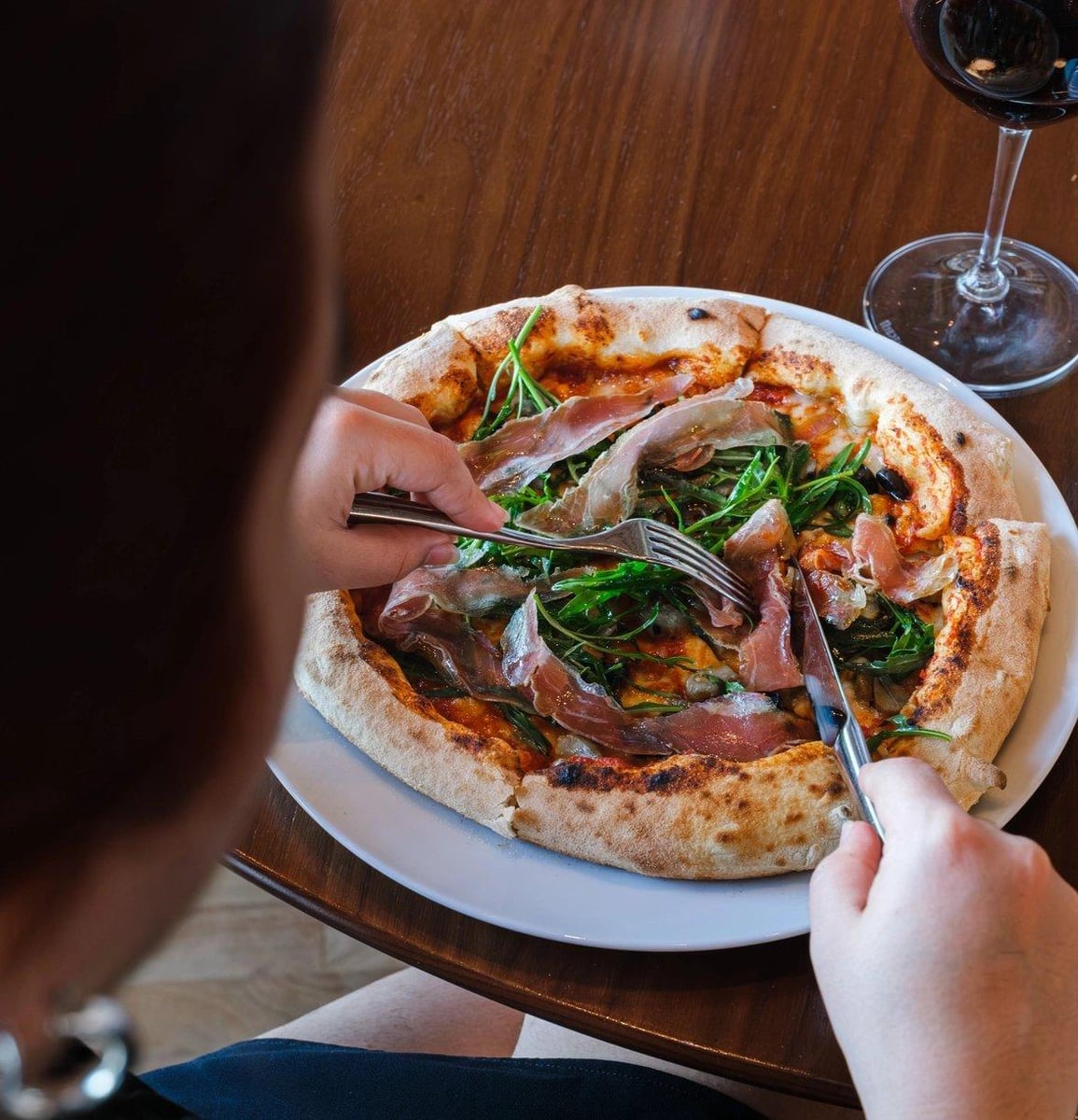 Hands cutting a pizza topped with cured ham and rocket, a glass of red wine beside