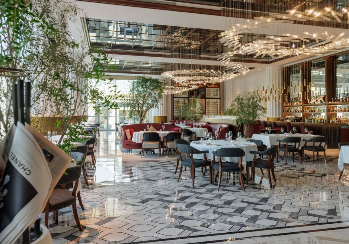 Tables laid in white beneath swirling light sculptures and a mirrored ceiling, olive trees along the windows and red banquettes beyond