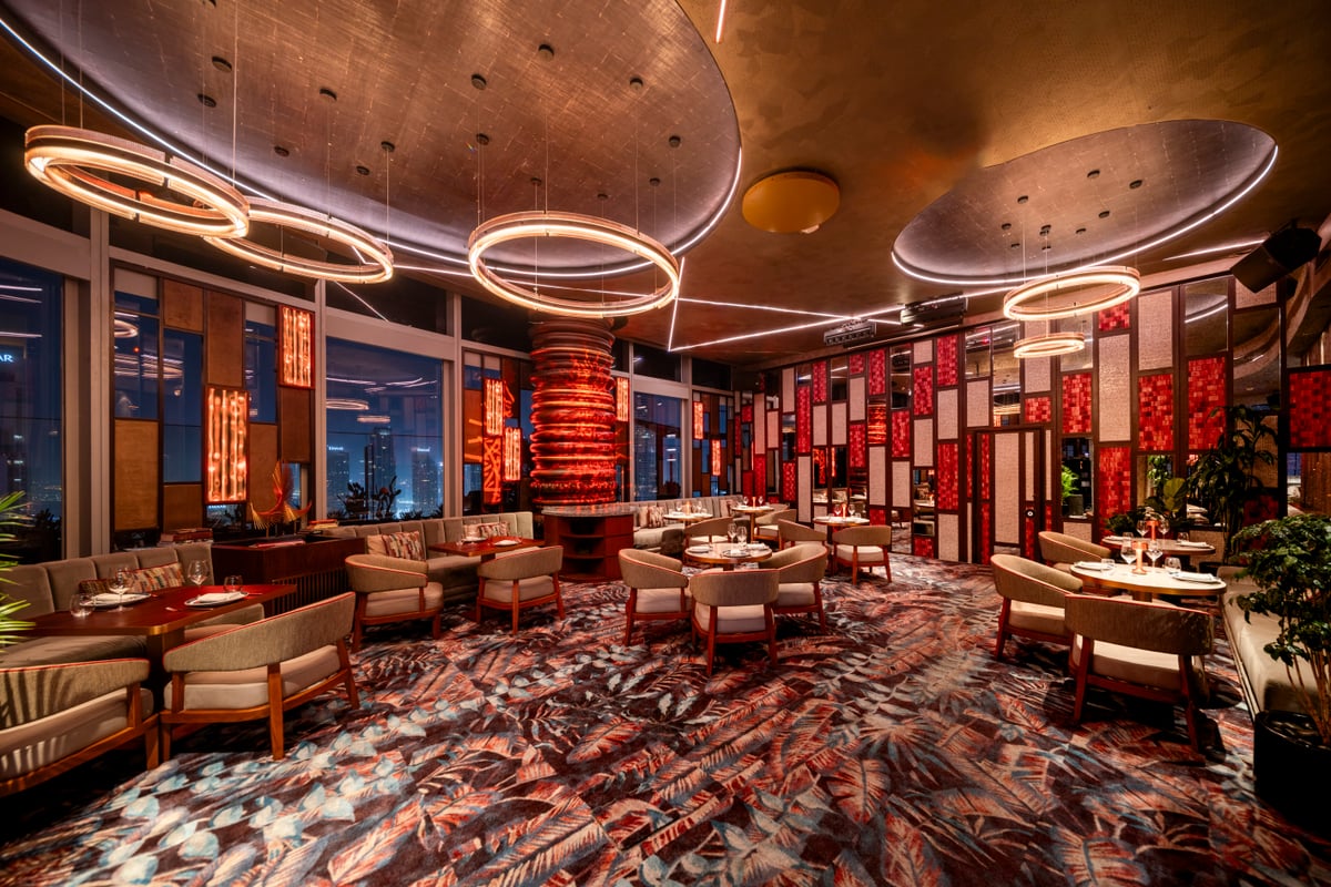 A dining room of red lacquered panels, ring-shaped pendant lights and a patterned carpet, city lights outside
