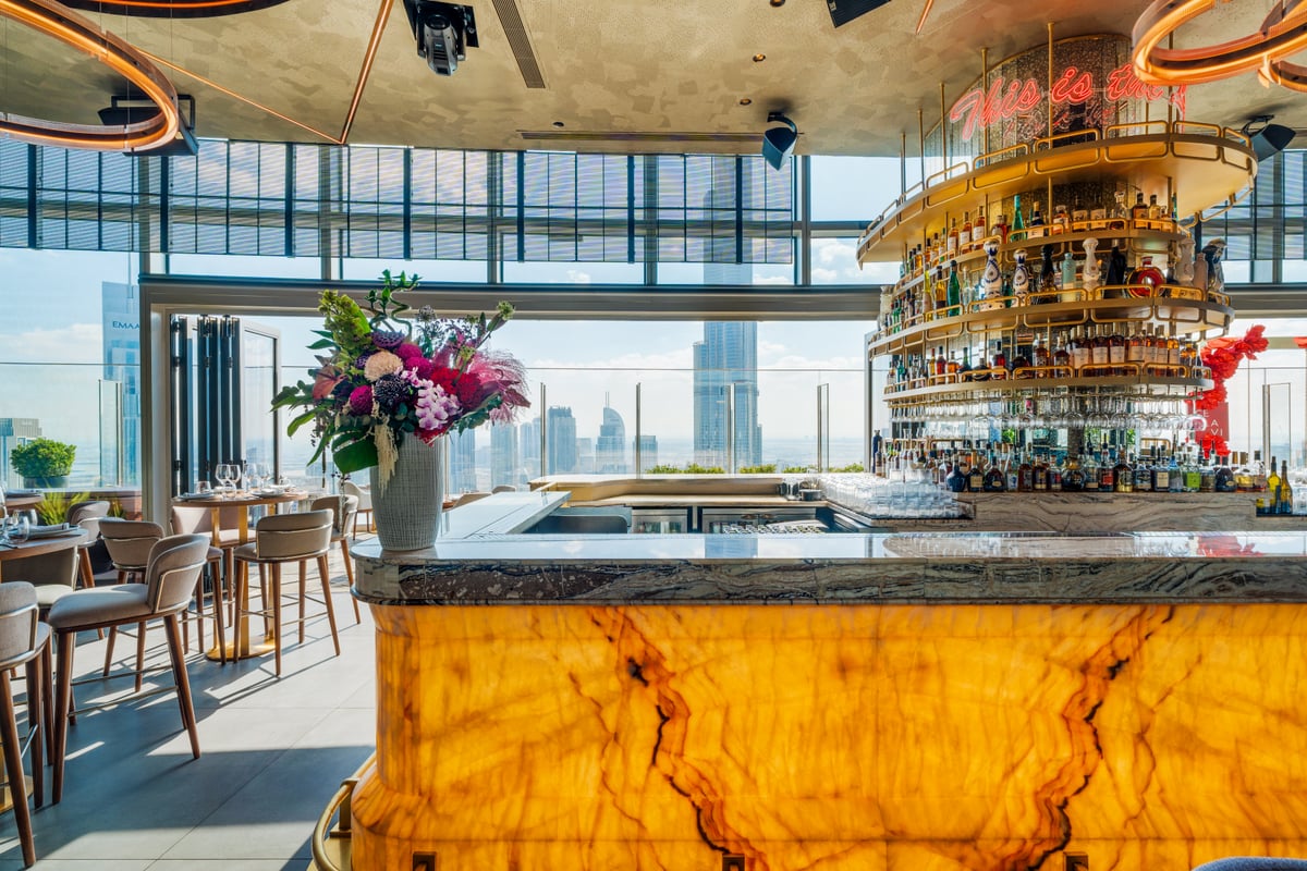 A glowing amber onyx bar topped with flowers, a tiered bottle display behind and the skyline through tall windows