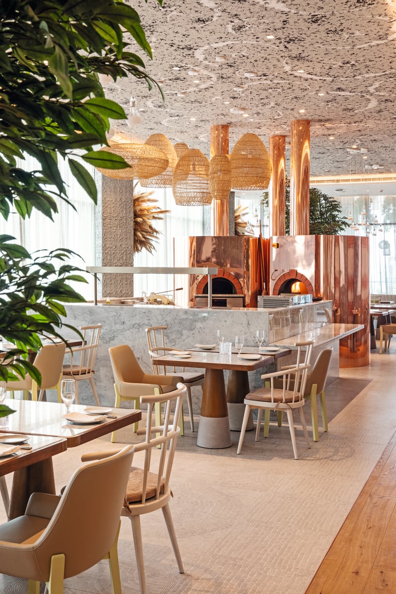 Pale wooden tables and cream chairs beneath a textured ceiling and rattan pendants, the copper pizza oven at the back