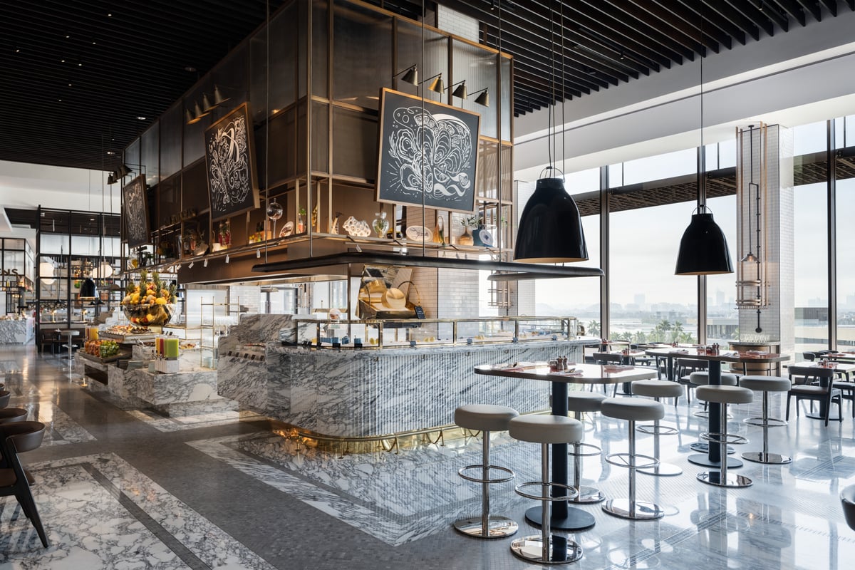 A marble-clad food counter beneath a dark steel frame and black pendant lamps, high stools and tables by tall windows