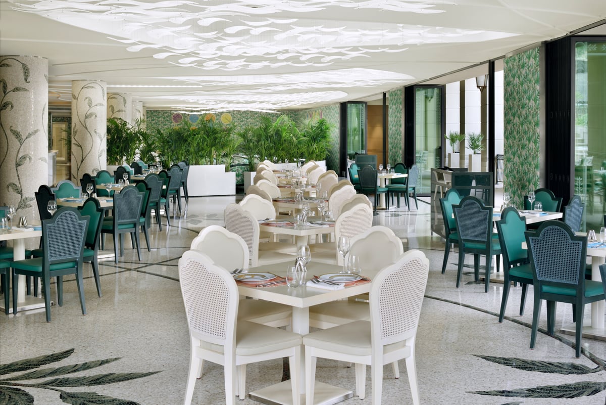 White and green chairs at long tables under a sculpted white ceiling, mosaic columns and greenery along the windows