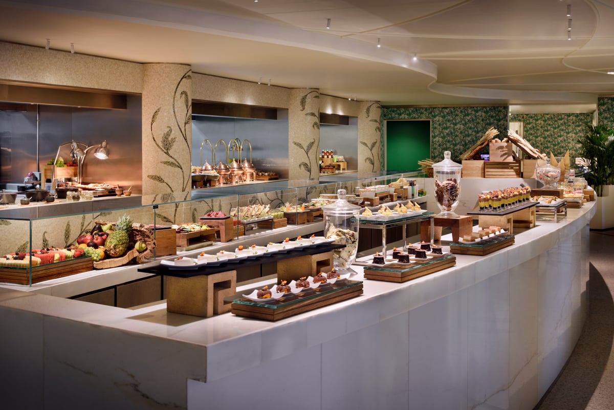 A long curved buffet counter of pastries, fruit and desserts beneath botanical mosaic walls