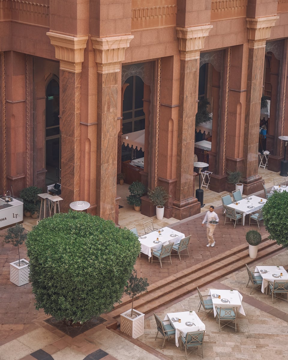 White-clothed tables on a courtyard terrace among Emirates Palace's rose-colored columns, a clipped shrub in the center