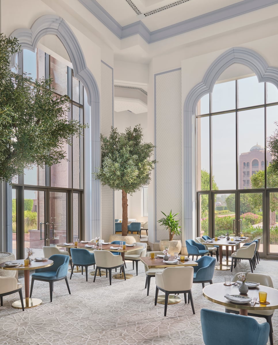Tall arched windows and olive trees in a bright, high-ceilinged dining room of blue and cream chairs