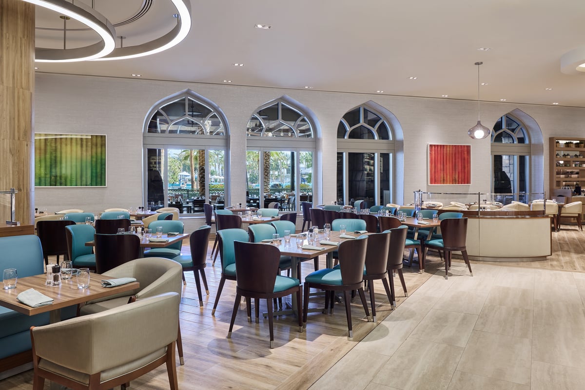 Rows of tables with teal and gray chairs beneath tall arched windows, bright artwork on the white walls