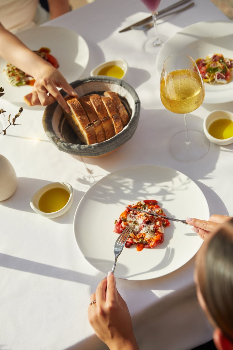 Hands reaching for grilled bread and a tomato dish across a white tablecloth with olive oil and white wine