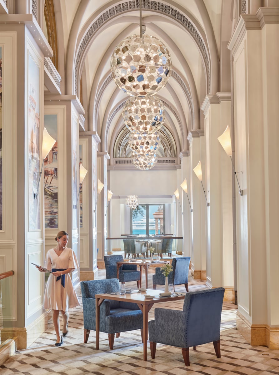 Blue velvet armchairs along a vaulted gallery hung with globe chandeliers, a host walking through