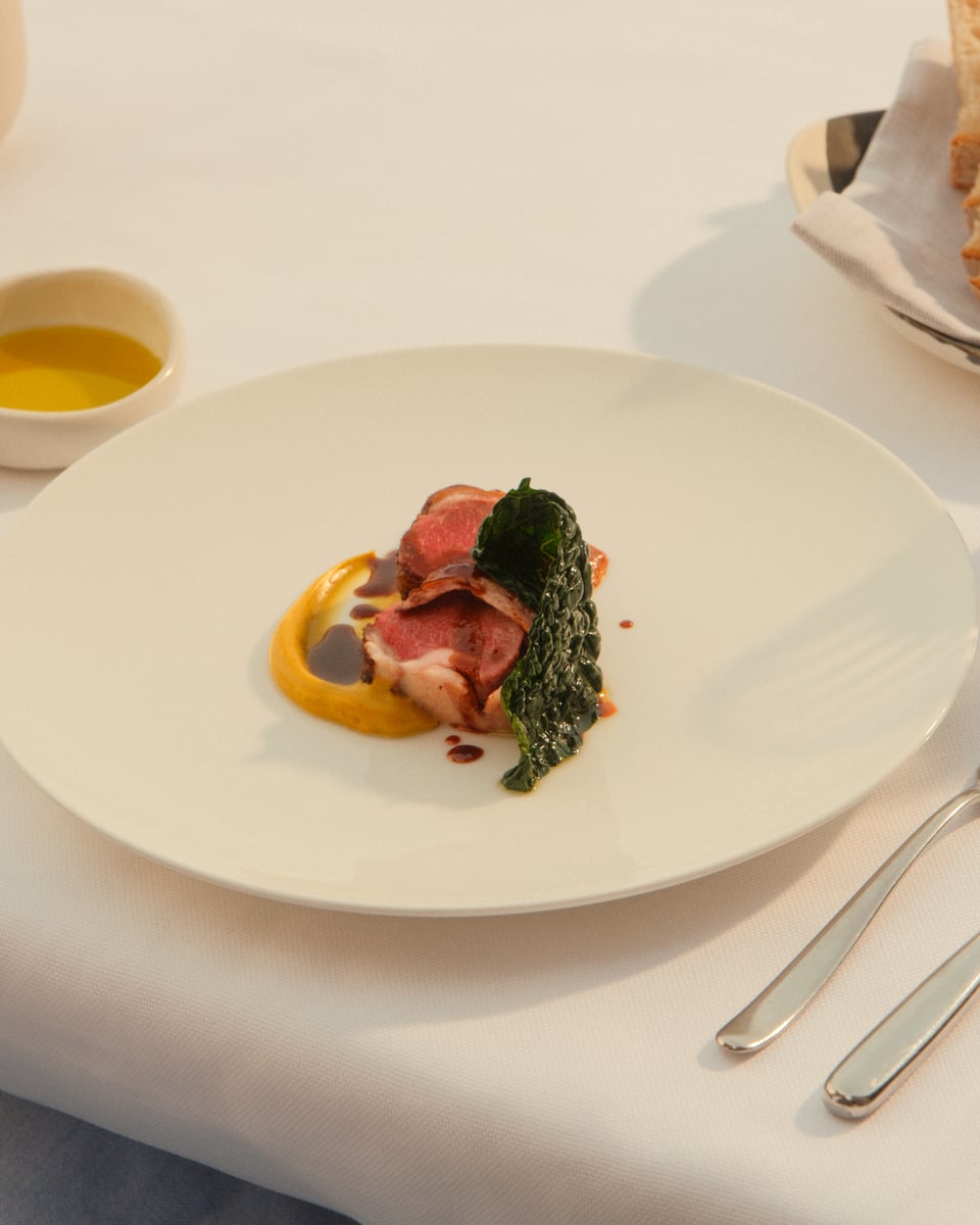 A slice of lamb with dark greens and a smear of orange purée on a wide white plate