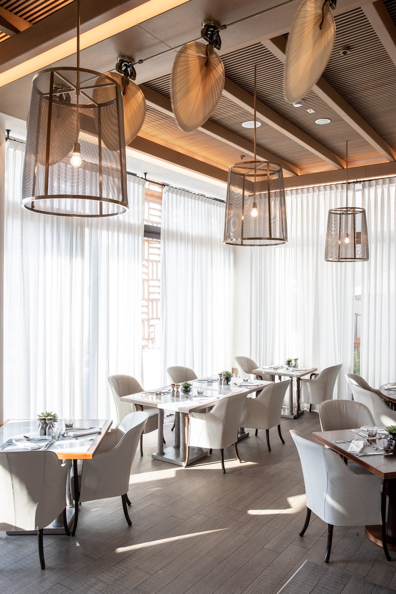 White upholstered chairs at laid tables under woven lantern pendants, sheer curtains drawn across tall windows