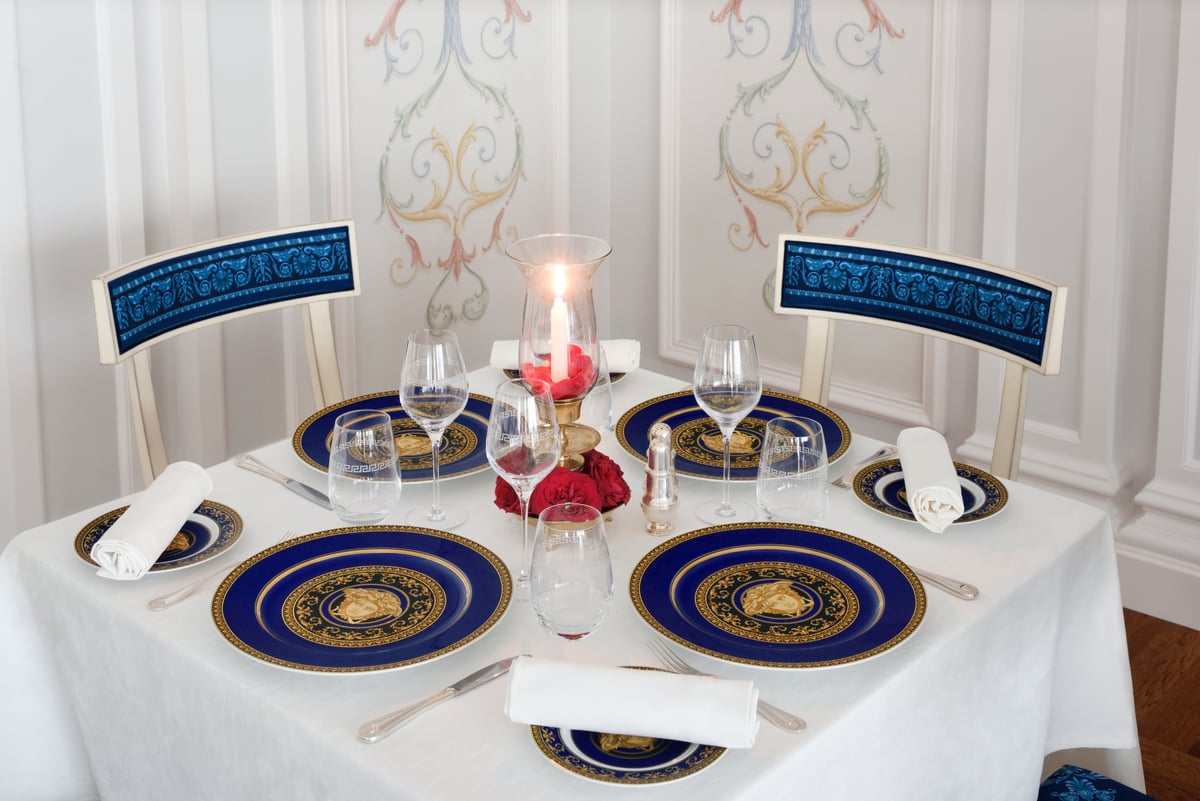 A table laid with blue-and-gold Versace plates, a candle and red roses between two blue-backed chairs