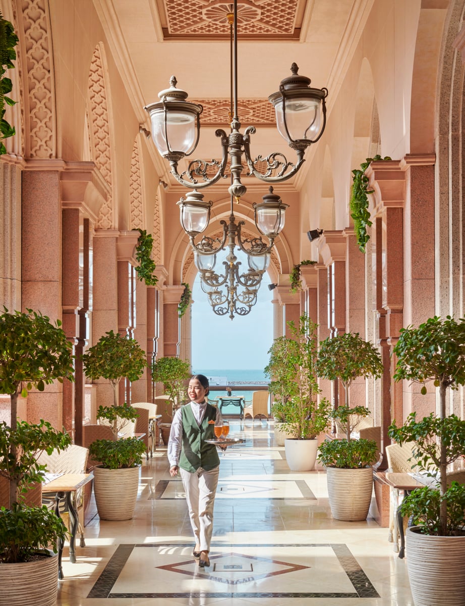 A long colonnade of pink marble arches with brass lanterns and potted trees, the sea at the far end